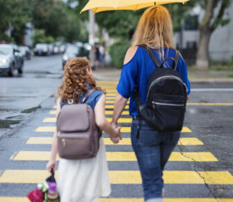 Young girl and her single mother walking to school. Back view of them holding hands, crossing city street on rainy day.