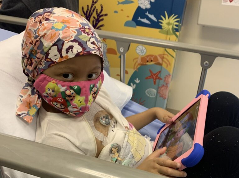 Girl in a hospital bed wearing a head scarf and face mask using a tablet computer.