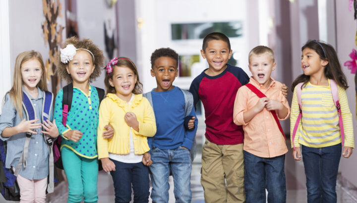 A group of seven children standing in a row in a school hallway, laughing and smiling at the camera. The little boys and girls are kindergarten or preschool age, 4 to 6 years.