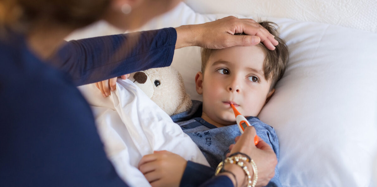 Sick boy with thermometer laying in bed and mother hand taking temperature. Mother checking temperature of her sick son who has thermometer in his mouth. Sick child with fever and illness while resting in bed.