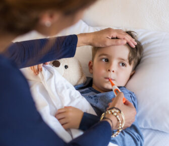 Sick boy with thermometer laying in bed and mother hand taking temperature. Mother checking temperature of her sick son who has thermometer in his mouth. Sick child with fever and illness while resting in bed.