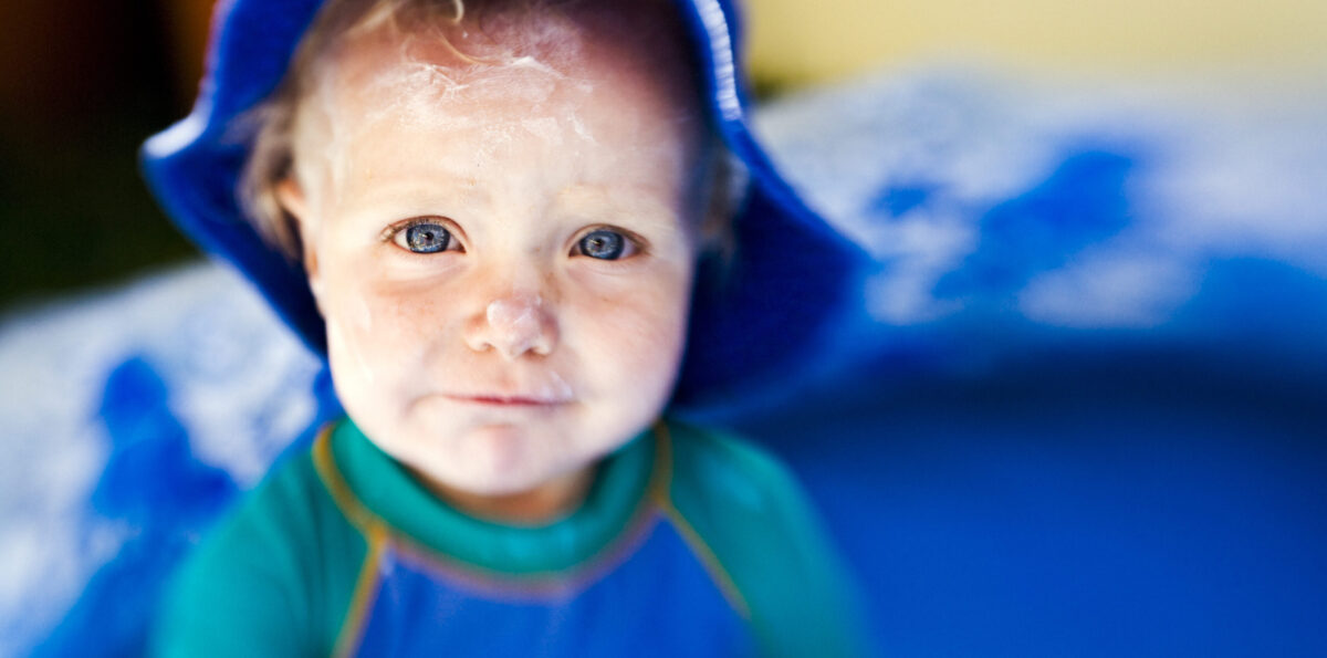 Year-old girl in a paddling pool, face covered in sunscreen and wearing a hat and anti-sunburn suit.