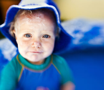 Year-old girl in a paddling pool, face covered in sunscreen and wearing a hat and anti-sunburn suit.