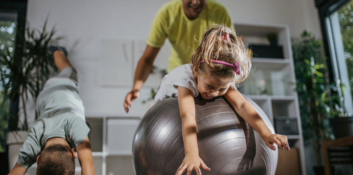 Child rolling on large workout ball with adult behind her and a young boy doing yoga next to them