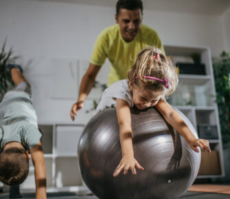 Child rolling on large workout ball with adult behind her and a young boy doing yoga next to them